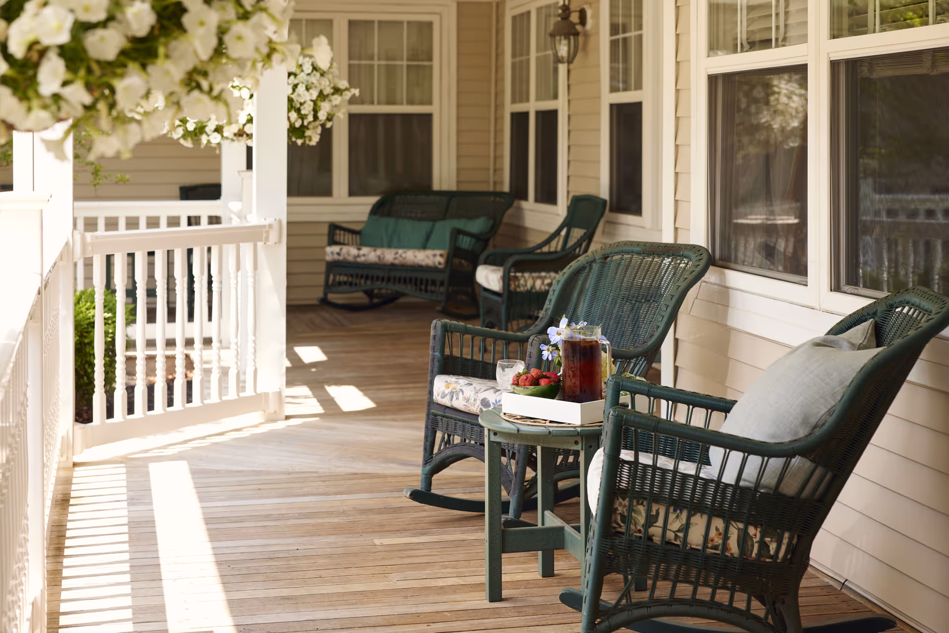 A sunlit wooden porch with green wicker rocking chairs and a small table holding a pitcher of iced tea, a glass with ice, and a bowl of strawberries. White flowers hang from above, and the porch is surrounded by white railings and beige siding with multiple windows.