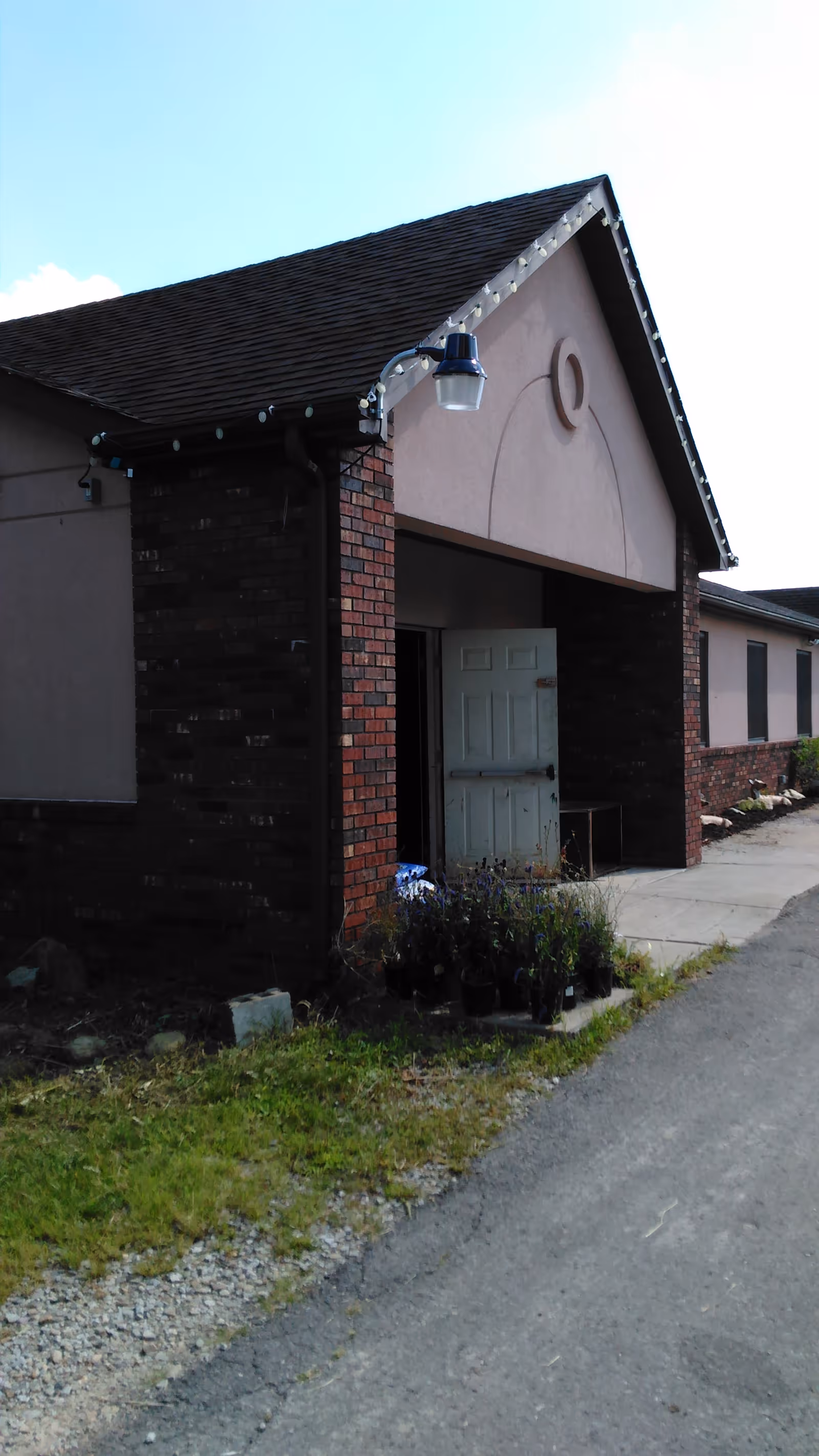 Exterior view of a building with a brick and stucco facade, featuring a peaked roof and an open door. There are several potted plants placed near the entrance along a paved walkway and gravel area.