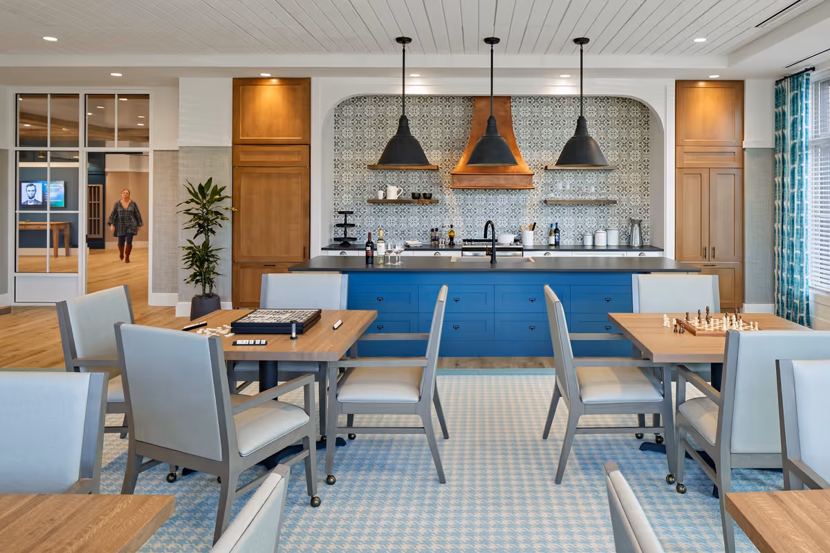 Bright communal dining and activity room with tables and chairs facing a blue kitchen island, pendant lights, and patterned tile backsplash.