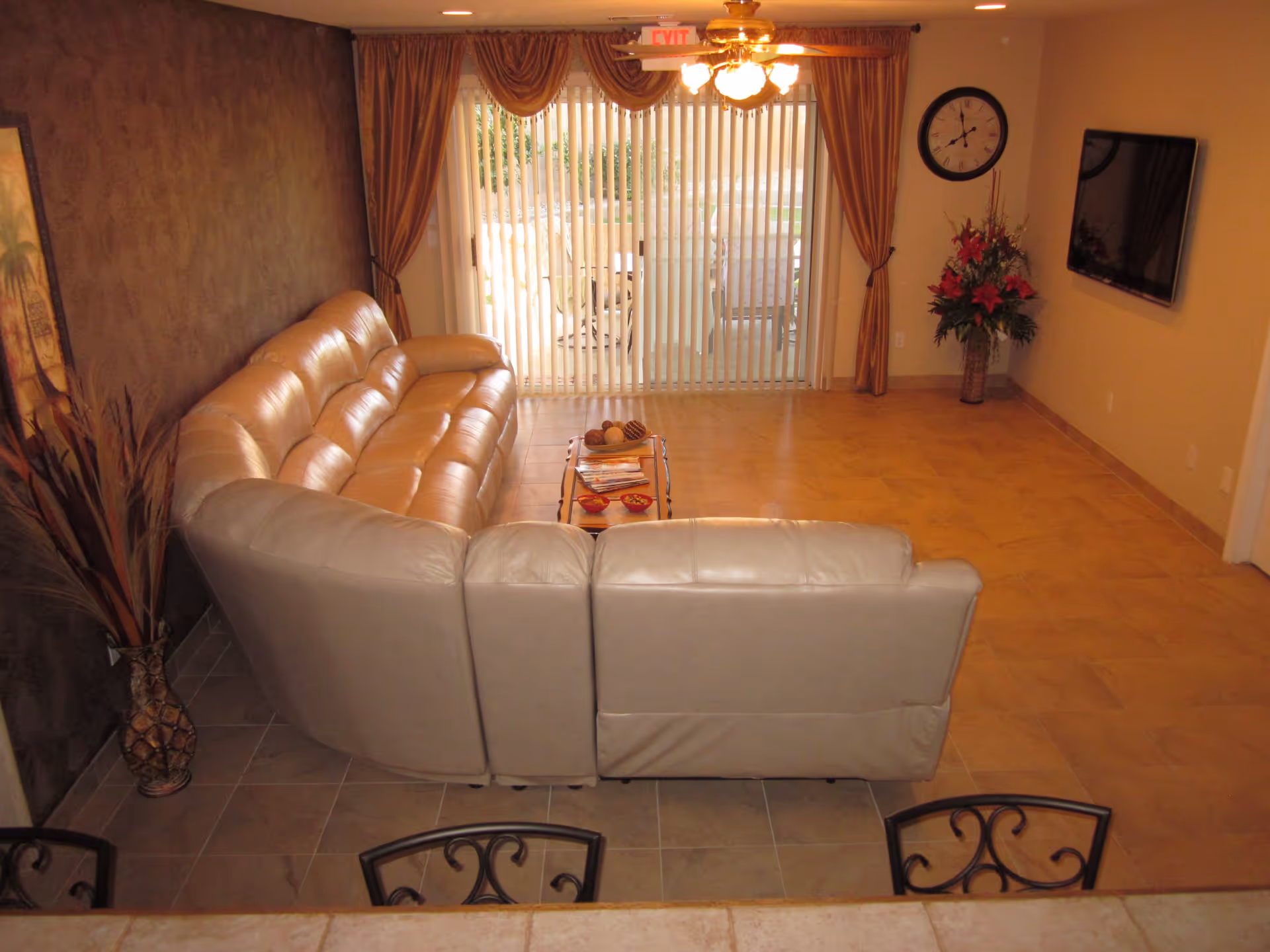 Interior view of a living room in Parkview Assisted Living featuring a large beige leather sectional sofa, a small coffee table with decorative items, a wall-mounted flat screen TV, a large clock, a vase with flowers, and vertical blinds covering a sliding glass door leading to an outdoor patio.