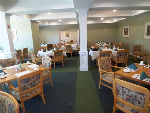 A dining room with multiple wooden tables and chairs arranged neatly. Each table is set with white tablecloths, napkins, cups, and silverware. The room has green walls, carpeted floors with a green runner, and framed artwork on the walls. The space is well-lit with ceiling lights.