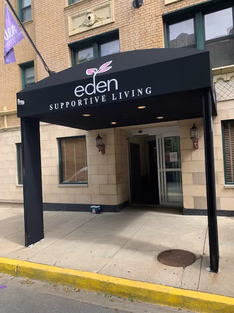 Entrance to Eden Supportive Living facility with a black awning displaying the facility's name and logo above a sidewalk and building entrance.