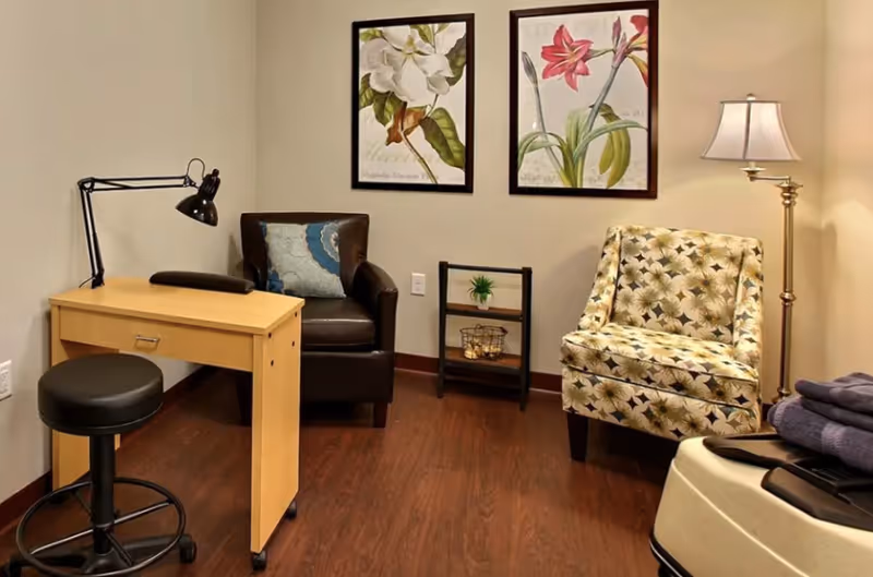 Cozy sitting room with two armchairs, a small desk and lamp, botanical wall art, a floor lamp, and a small shelving unit on hardwood flooring.