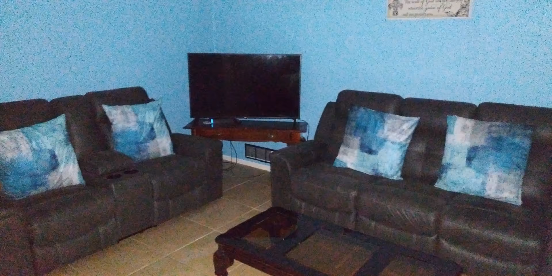 A living room with two dark brown reclining sofas, each adorned with blue and white patterned throw pillows. A flat-screen TV is placed on a wooden corner stand against a light blue wall. The floor is tiled, and a dark wooden coffee table is positioned in front of the sofas.