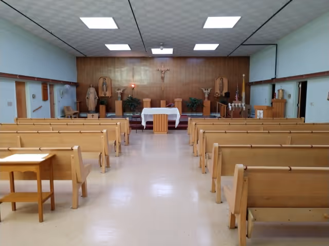 Interior view of a small chapel with wooden pews arranged in rows facing an altar. The altar is covered with a white cloth and has religious statues and a crucifix on the wooden-paneled wall behind it. The ceiling has recessed lighting panels.
