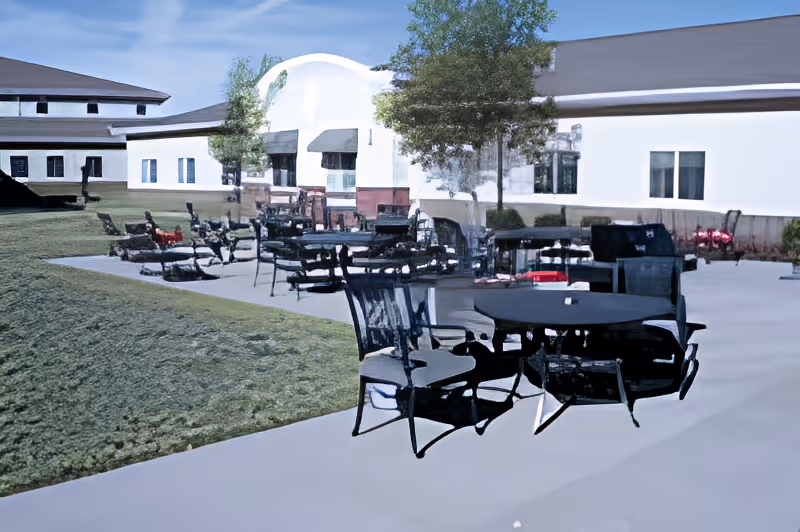 Outdoor patio with round tables and chairs on a concrete area beside a single-story white building and lawn.