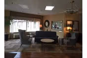 A cozy living room area with a dark sofa, two gray armchairs, a round coffee table, and a side table with a lamp. The room has large windows with curtains, a mirror on the wall, and a ceiling light fixture. The floor is carpeted with a patterned rug.