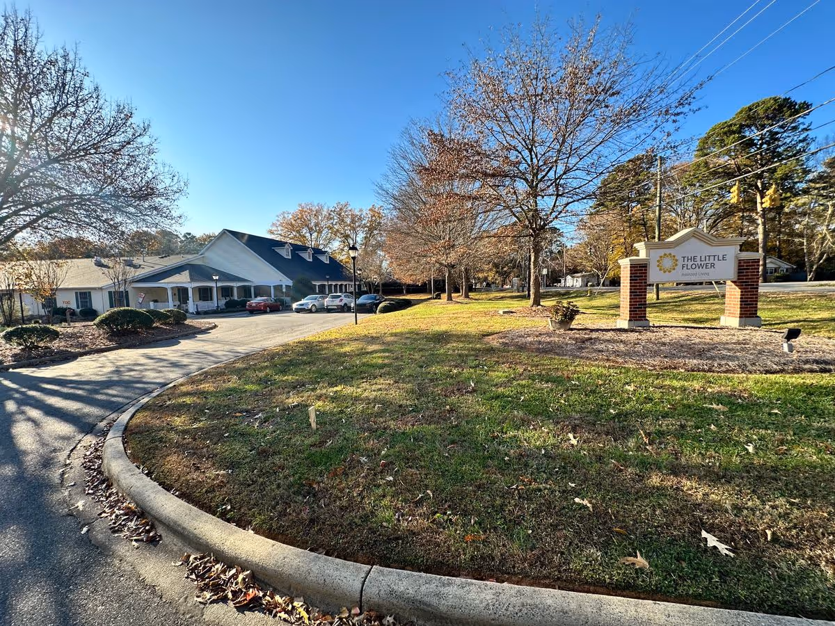 Exterior front view of The Little Flower assisted living facility with a driveway, parked cars, lawn, trees, and a sign reading 'The Little Flower'.