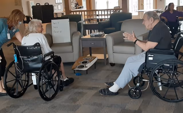 Two people in wheelchairs sit facing each other in a lounge area playing a tabletop ring-toss game with armchairs and a small table behind them.