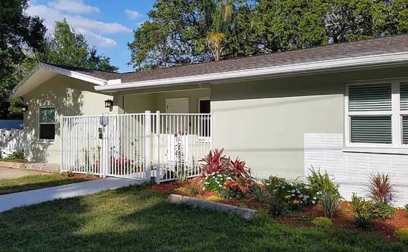 Exterior view of a single-story building with light green walls, white trim, and a white fence enclosing a small garden area with various plants and flowers. The building is surrounded by green grass and trees under a blue sky with some clouds.