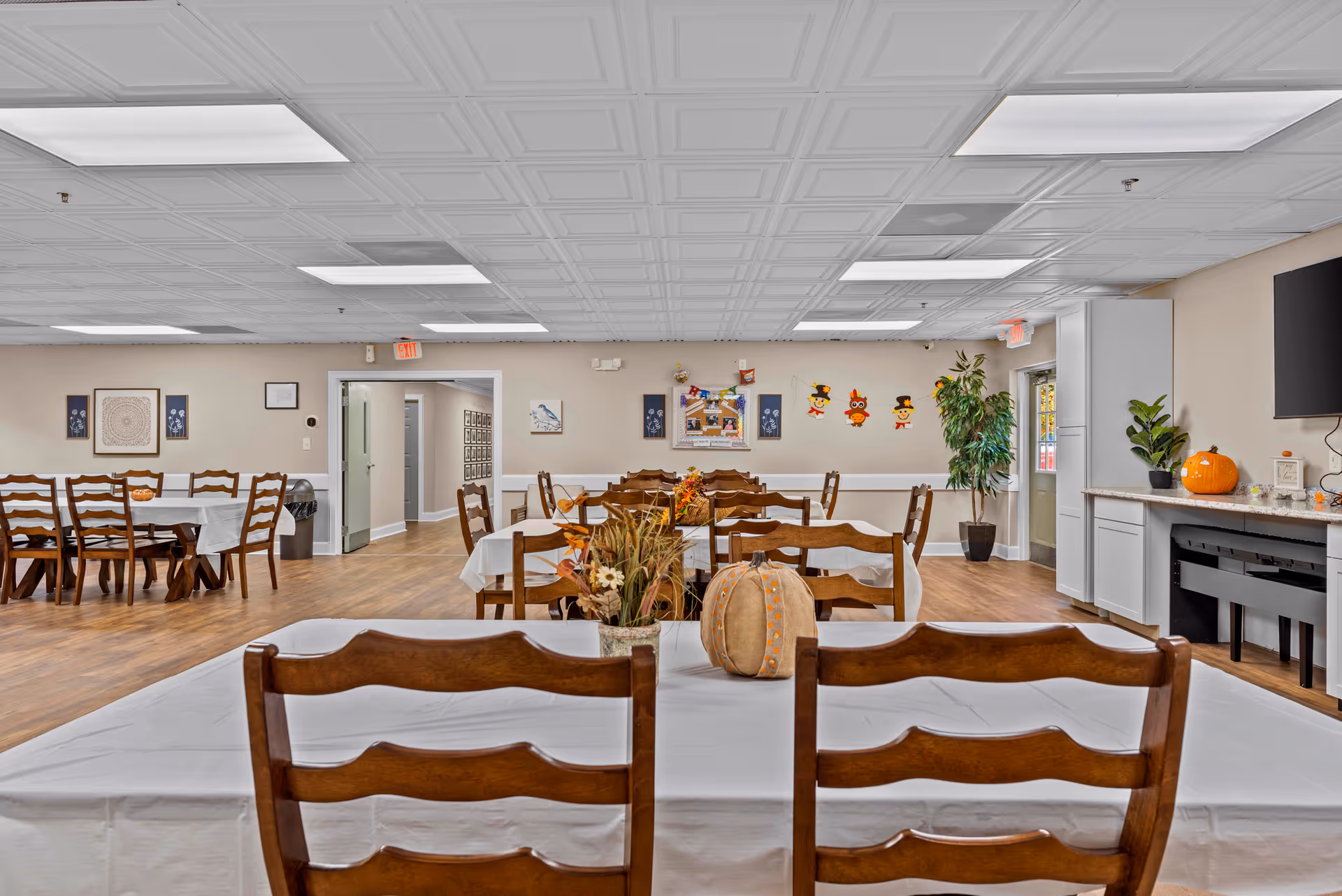 A spacious dining room with multiple wooden tables and chairs arranged neatly. The tables are covered with white tablecloths and decorated with autumn-themed centerpieces including pumpkins and flowers. The room has a white paneled ceiling with fluorescent lighting, beige walls adorned with framed artwork and seasonal decorations, and a wooden floor. There is a doorway leading to another room and a TV mounted on the wall to the right.