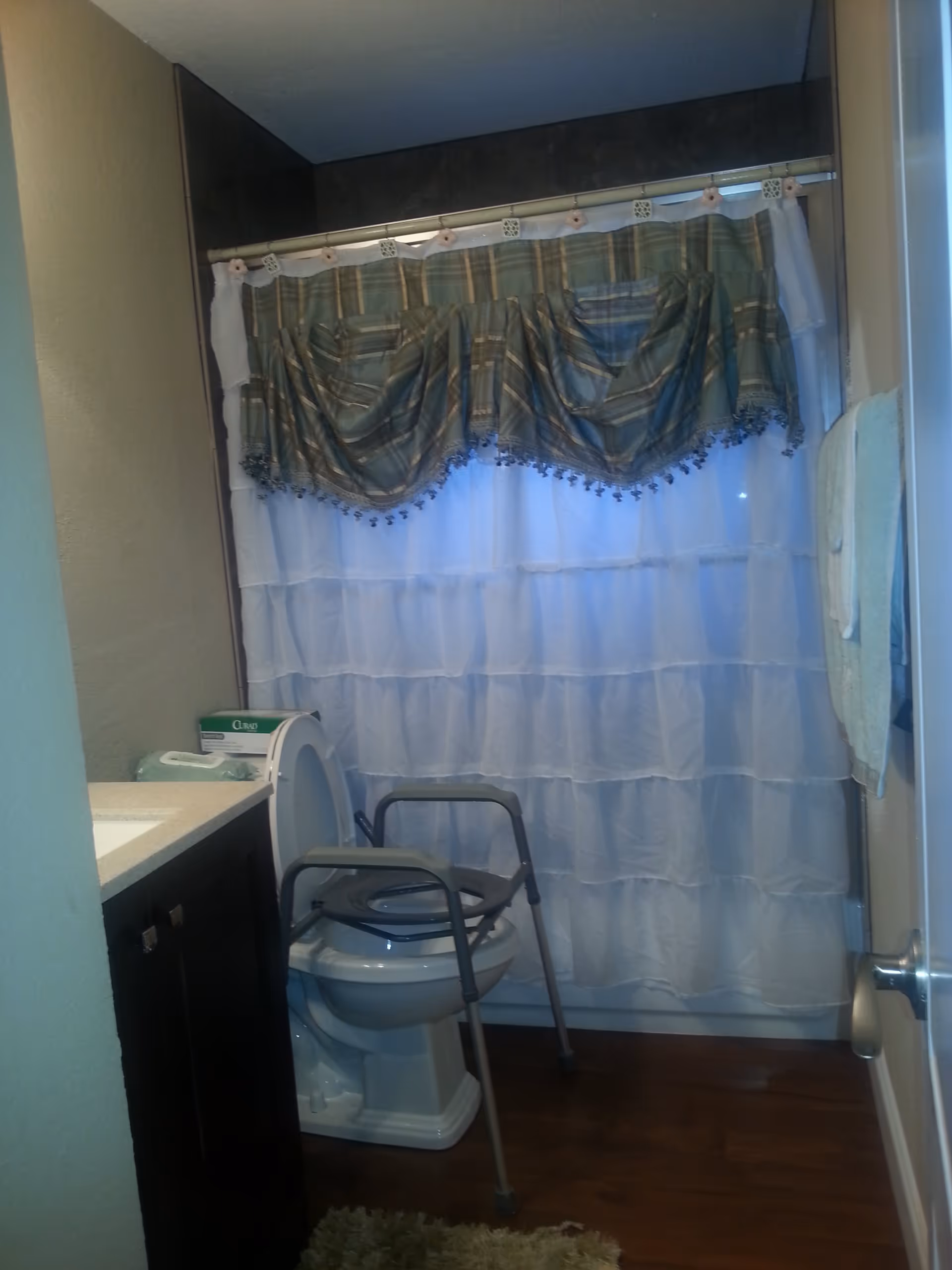 Bathroom with a toilet fitted with a metal commode frame, ruffled shower curtains with a valance, a vanity and a towel on the wall.