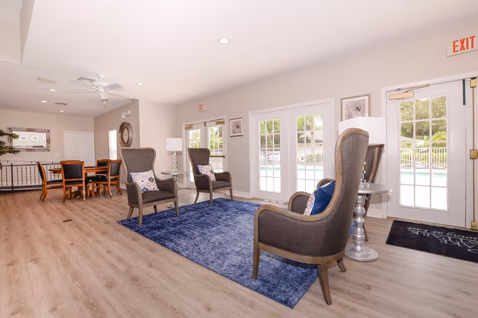 A bright and spacious common area with three high-back armchairs arranged around a blue rug, each with a side table and lamp. Behind the seating area are large glass doors leading outside to a pool. In the background, there is a dining table with chairs and a large wall clock. The room has light wood flooring and neutral-colored walls.