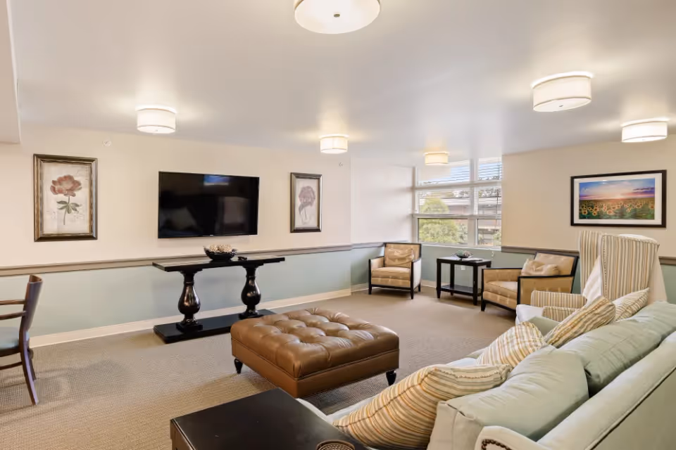 A well-lit living room with a large light blue sofa adorned with striped pillows, a brown tufted ottoman in the center, two beige armchairs near a window, a flat-screen TV mounted on the wall, and framed floral artwork on the walls.