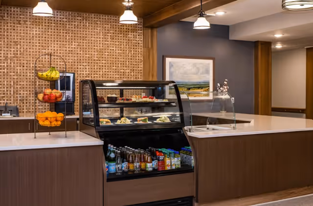 Snack bar with a glass display case of sandwiches and drinks, a three-tier fruit stand, and a countertop in a communal dining area.