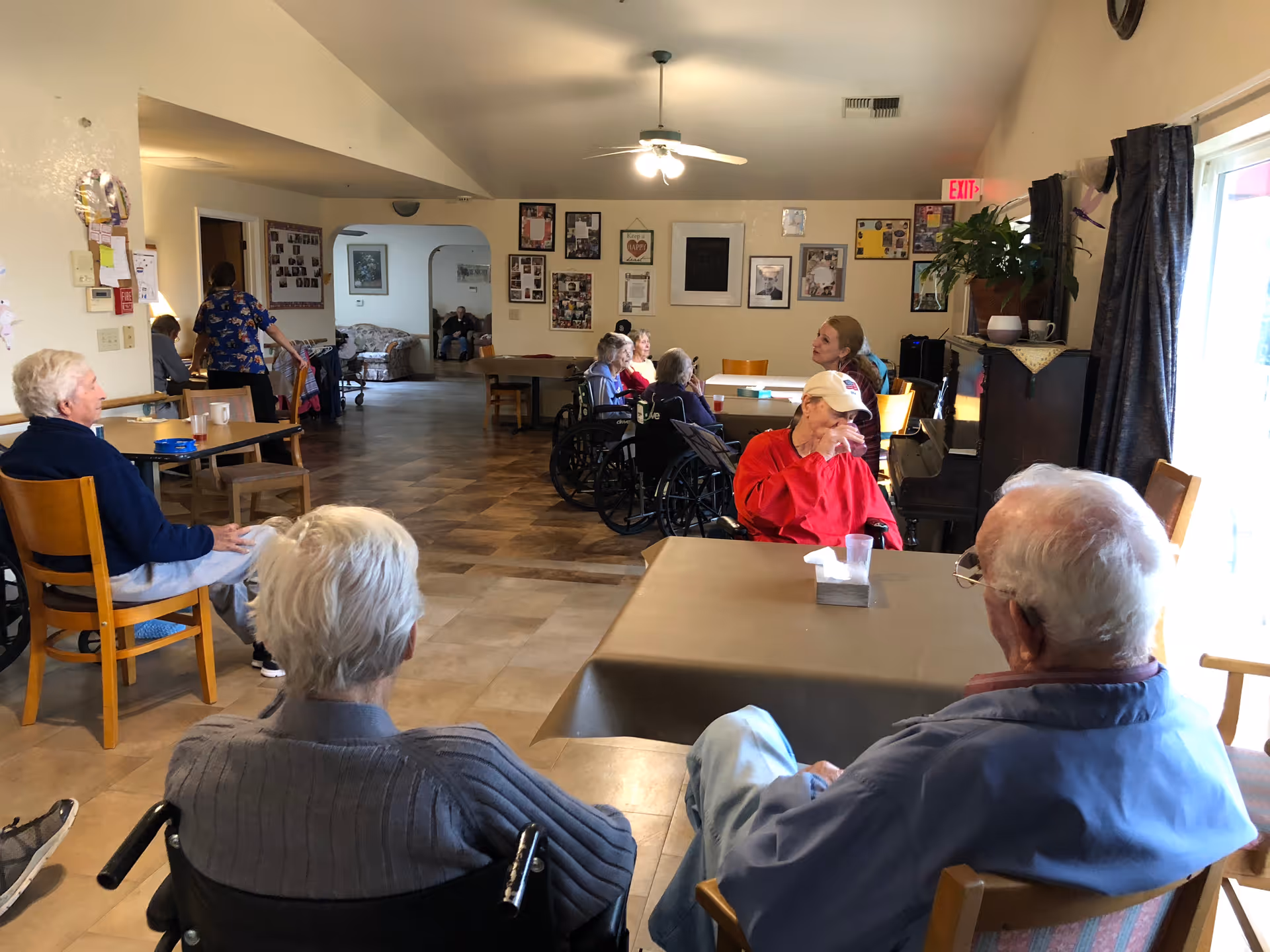 A group of elderly people sitting in a common area of an assisted living facility. Some are seated in wheelchairs while others are in regular chairs around tables. The room has a ceiling fan, framed pictures on the walls, and a large window with curtains letting in natural light. A staff member is seen walking in the background.