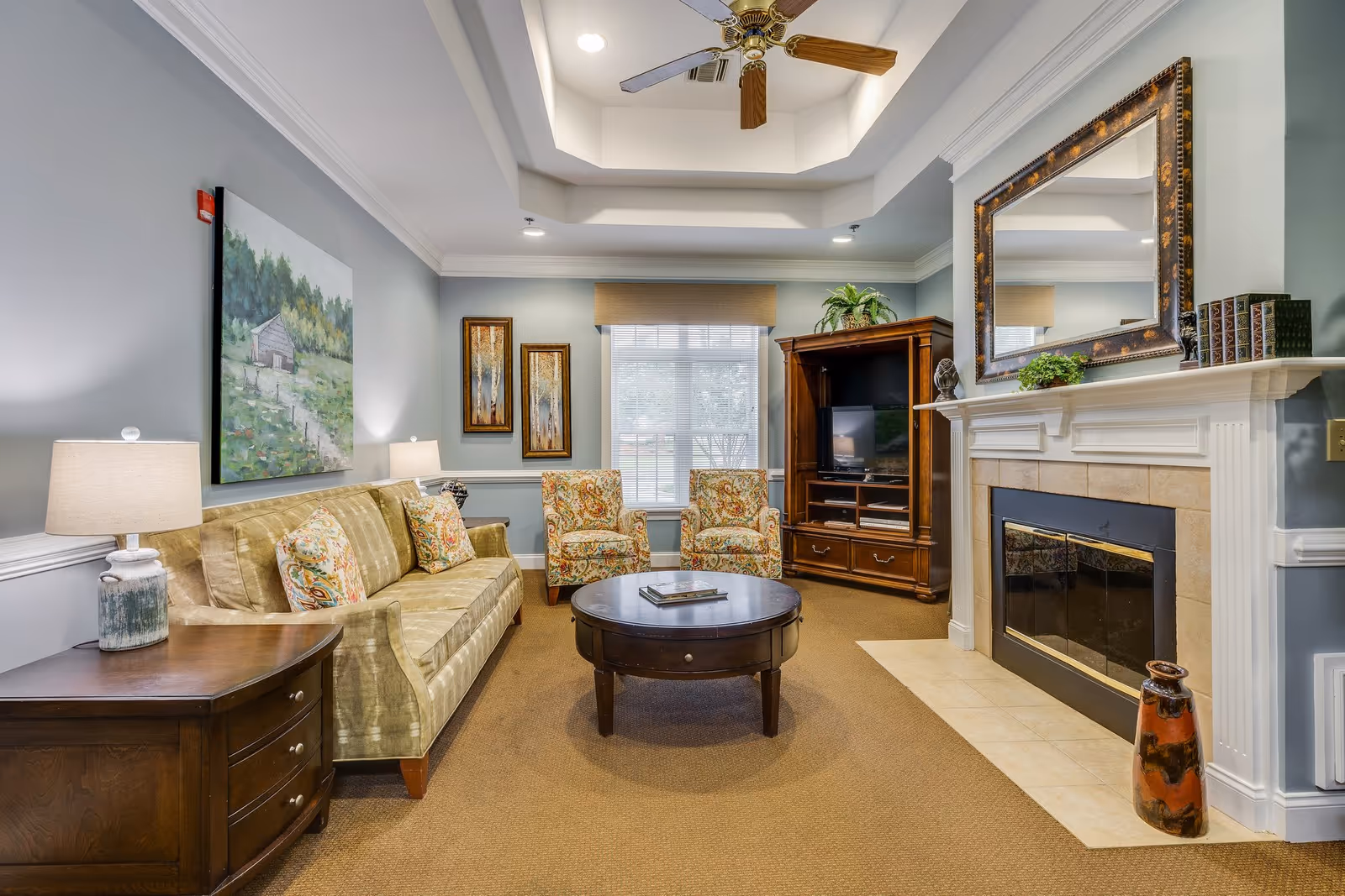 A cozy living room with a beige sofa adorned with patterned cushions, two matching armchairs, a round wooden coffee table, a wooden TV cabinet with a television, and a fireplace with a large decorative mirror above it. The walls are painted light blue and decorated with framed artwork. The room has a ceiling fan and a window with blinds letting in natural light.
