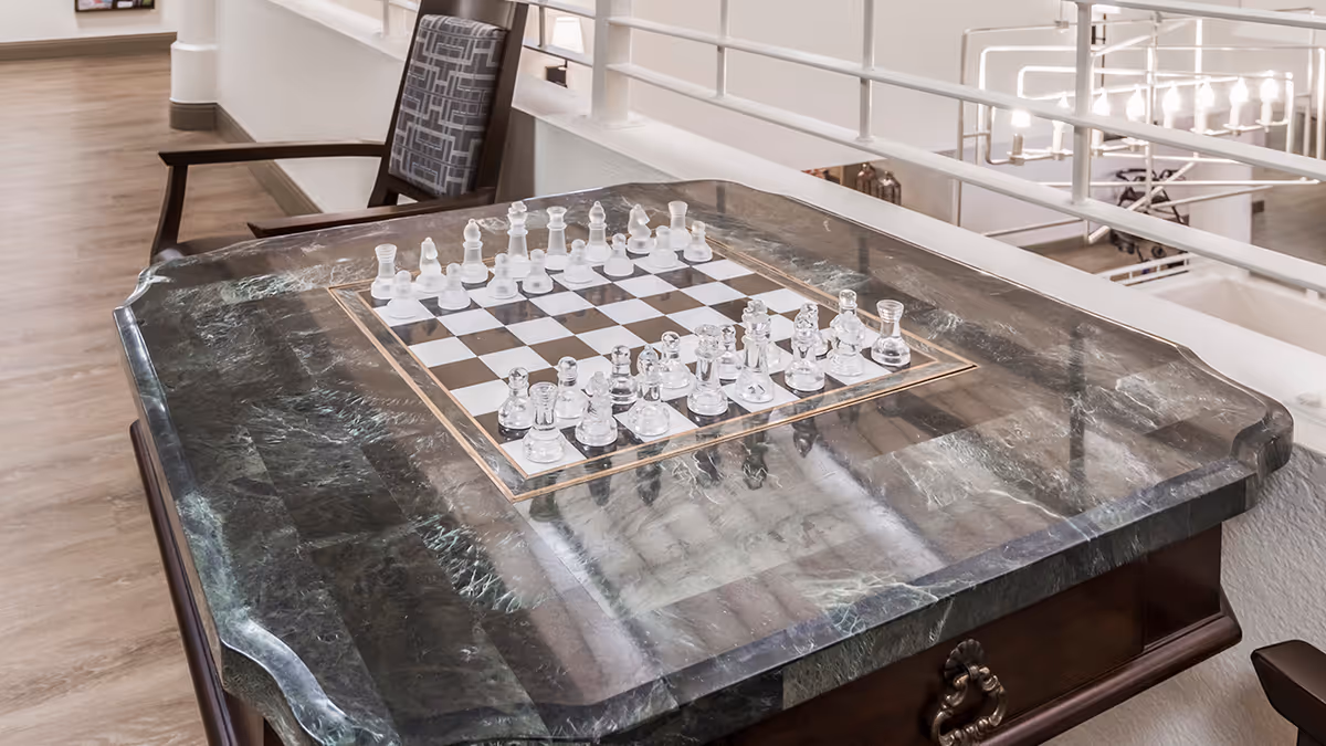 A polished marble chess table with a glass chess set arranged for play, situated in a well-lit indoor area with wooden flooring and a patterned chair nearby.