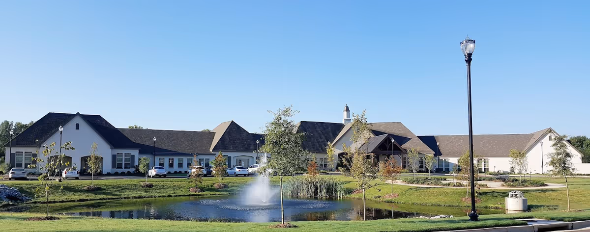 Exterior front view of a long single-story senior living building with a pond and fountain in front under a clear blue sky.