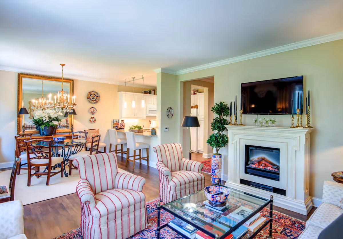 A bright and cozy living room with two striped armchairs, a glass coffee table with books and a decorative bowl, a fireplace with candles and a TV mounted above it. The room opens into a dining area with a wooden table, chairs, a chandelier, and a large mirror on the wall. The kitchen with white cabinets and a breakfast bar with stools is visible in the background.
