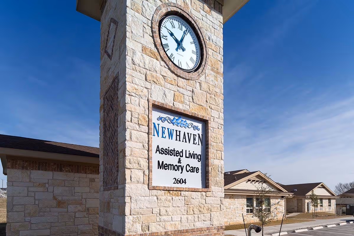 Stone clock tower with a large round clock and a sign that reads 'New Haven Assisted Living & Memory Care 2604' in front of a single-story building under a clear blue sky.
