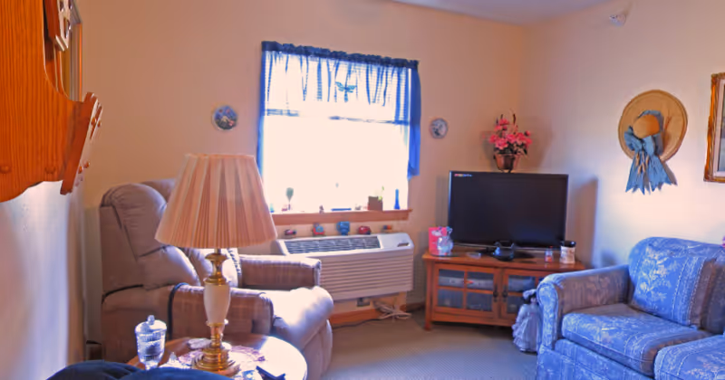 A cozy living room with a beige recliner chair, a blue patterned sofa, a wooden TV stand with a flat-screen TV, a table lamp on a side table, and a window with blue curtains letting in natural light. The walls are decorated with a straw hat with a blue ribbon and a framed picture.