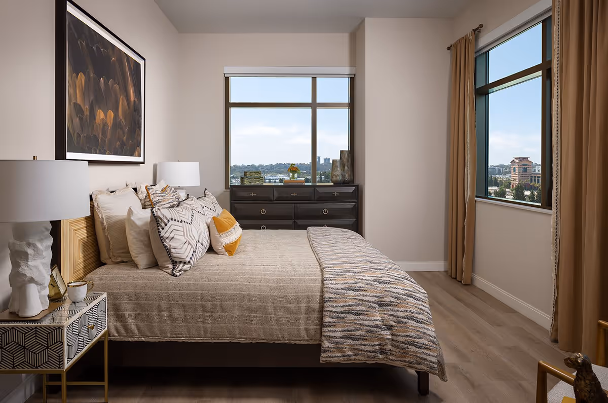 A well-lit bedroom with a large bed featuring multiple decorative pillows and a textured blanket. There are two windows with views of buildings and greenery outside. The room includes a dark dresser under one window, a patterned nightstand with a white lamp, and beige curtains on the other window. A framed abstract artwork hangs above the bed.