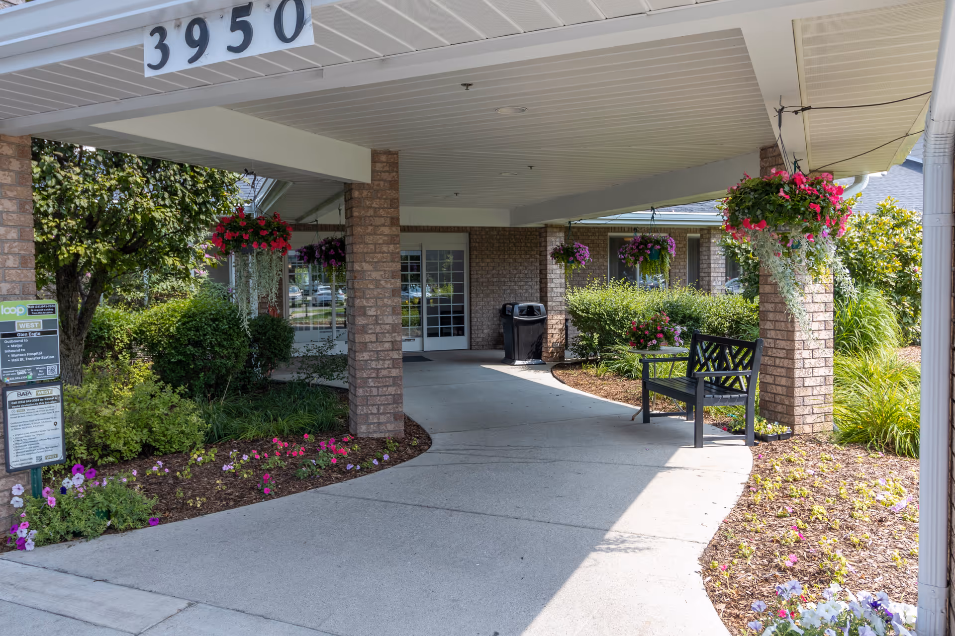 Covered entrance area of a senior living facility with brick pillars, hanging flower baskets, a black bench, and landscaped flower beds. The building number 3950 is displayed above the entrance, and there is a bus stop sign on the left side near greenery.