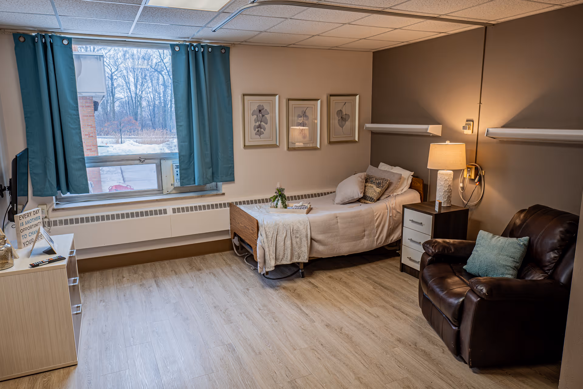 A cozy senior living bedroom featuring a single bed with pillows and a blanket, a bedside table with a lamp, a comfortable leather armchair with a cushion, a window with teal curtains, and framed botanical artwork on the wall. The room has wood flooring and a small dresser with a TV on top.