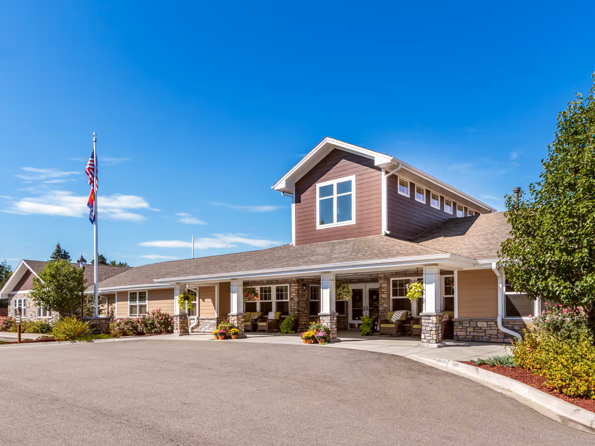 Exterior view of a single-story senior living facility building with a covered entrance, stone pillars, and seating areas. The building has beige and brown siding with white trim, surrounded by landscaped greenery and flowers under a clear blue sky. Two flagpoles with flags are visible on the left side.