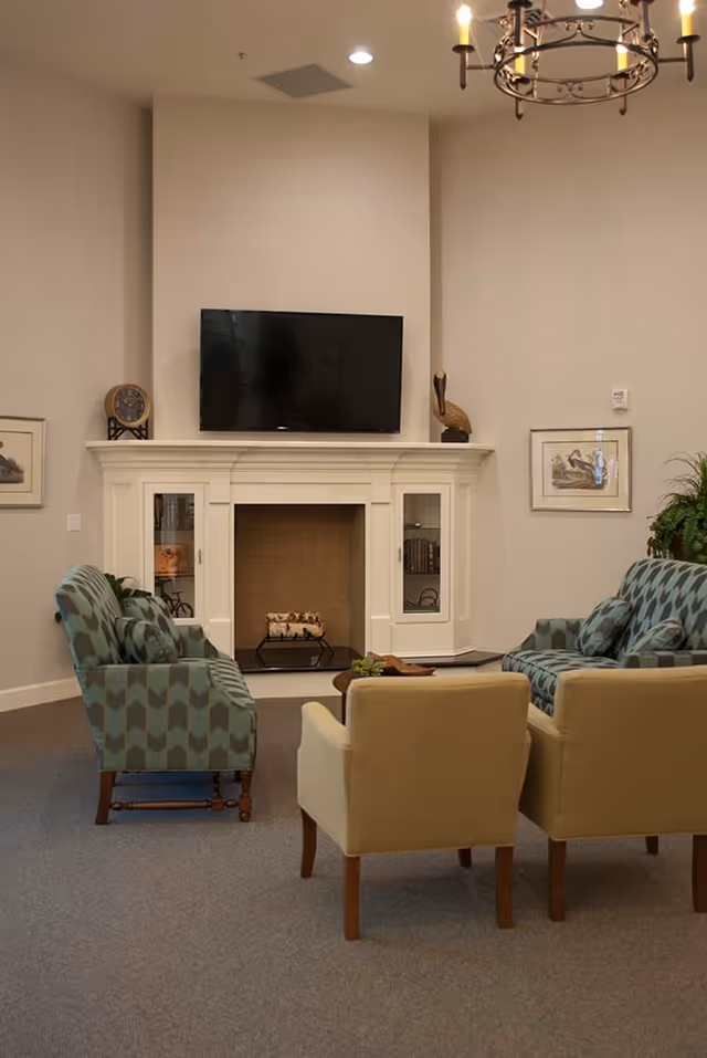 A cozy living room area with a white fireplace and a mounted flat-screen TV above it. The room features two patterned armchairs and two beige armchairs arranged around a small coffee table. Decorative items include framed artwork on the walls, a clock, a bird sculpture on the mantel, and a chandelier hanging from the ceiling.