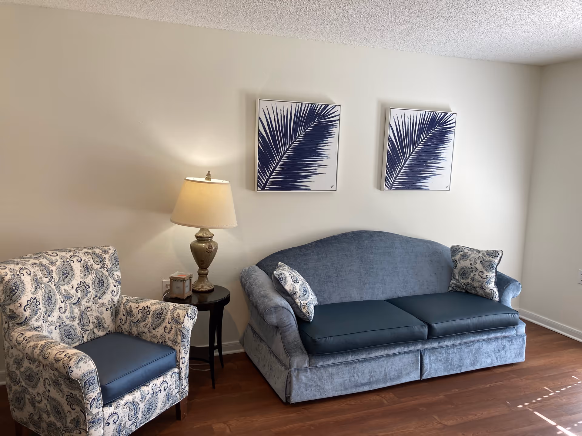 A cozy living room area featuring a blue upholstered sofa with two patterned cushions, a matching patterned armchair with a blue seat cushion, a small round black side table with a decorative lamp and a small item on it, and two framed artworks of blue palm leaves hanging on a white wall.