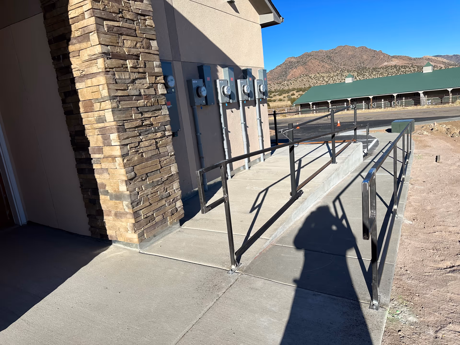 Concrete accessibility ramp with metal handrails beside a stone-clad exterior wall and utility meters, with mountains in the background.