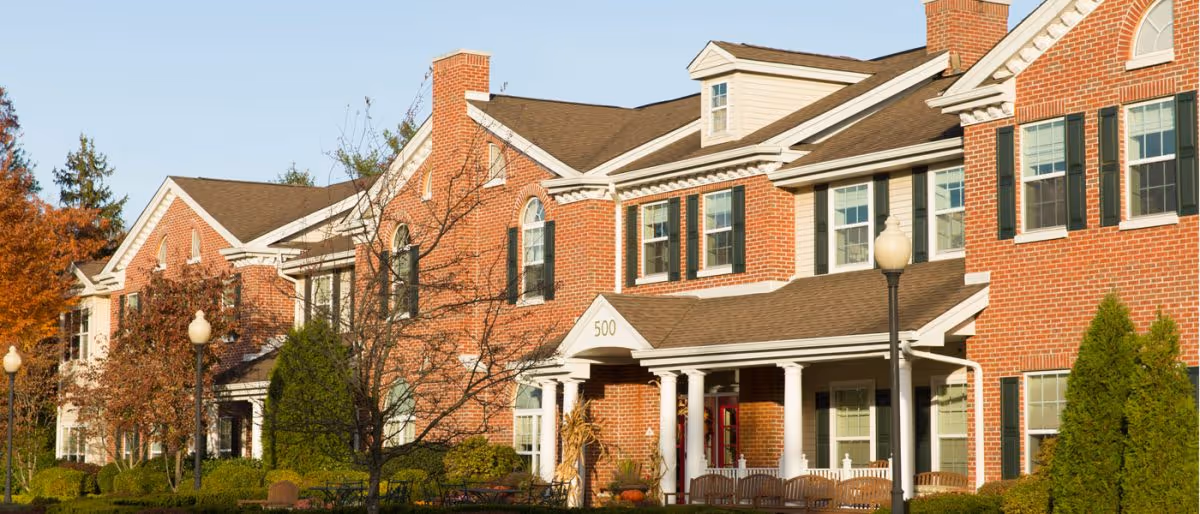 Exterior view of a large brick senior living facility building with multiple windows, white columns at the entrance, and a brown shingled roof. There are trees with autumn foliage and green shrubs in front of the building, along with street lamps and benches near the entrance.