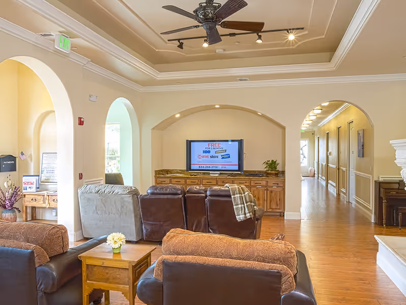A cozy living room area in a senior living facility with multiple comfortable chairs and sofas arranged around a television mounted in a recessed wall niche. The room features wooden flooring, a ceiling fan with lights, arched doorways, and a hallway leading to other rooms. There is a small wooden side table with a flower vase, and the space is warmly lit with natural and artificial light.