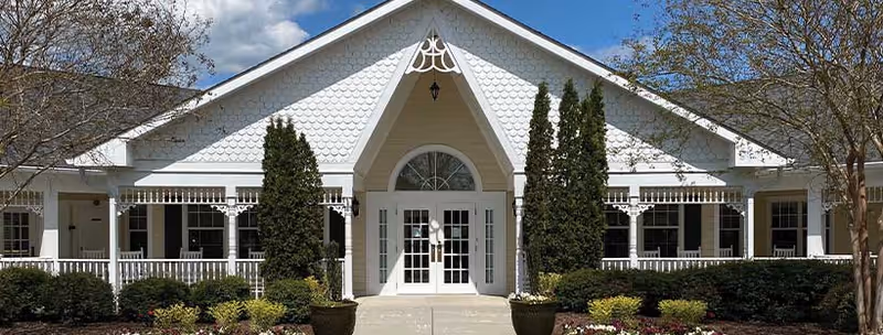 Front exterior view of a white building with a peaked roof, decorative trim, and large glass double doors at the entrance. The building is surrounded by neatly trimmed bushes, small trees, and landscaped flower beds under a partly cloudy blue sky.