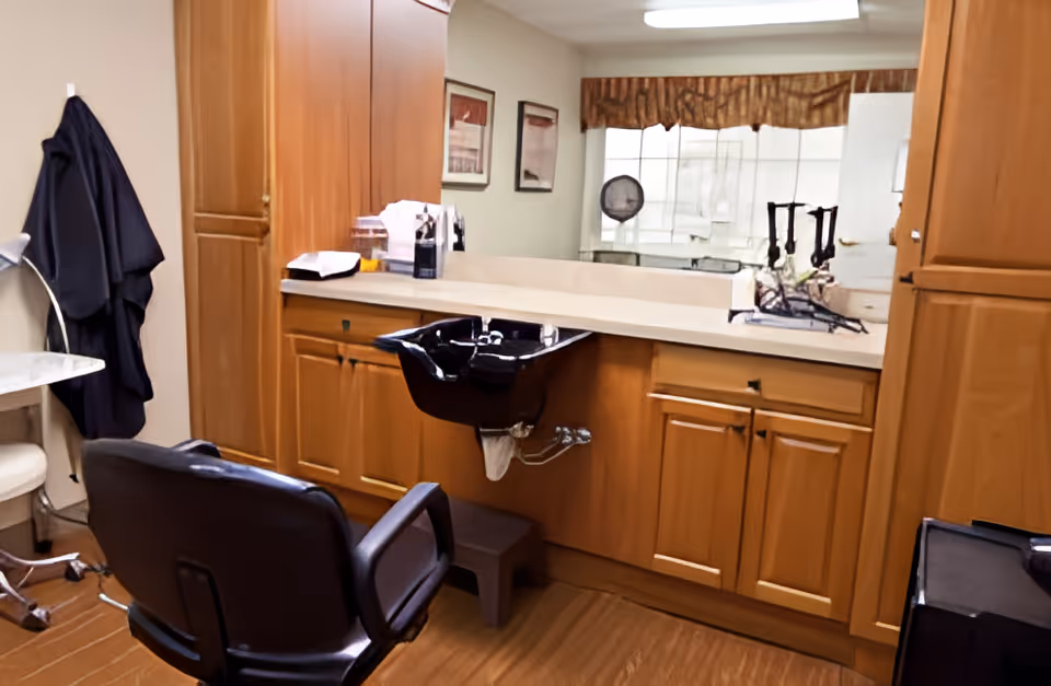 Interior view of a hair salon area in a senior living facility with a black salon chair facing a black wash basin. The room has wooden cabinets, a large mirror, and a window with a valance. A black cape hangs on the wall and there is a small stool under the basin.