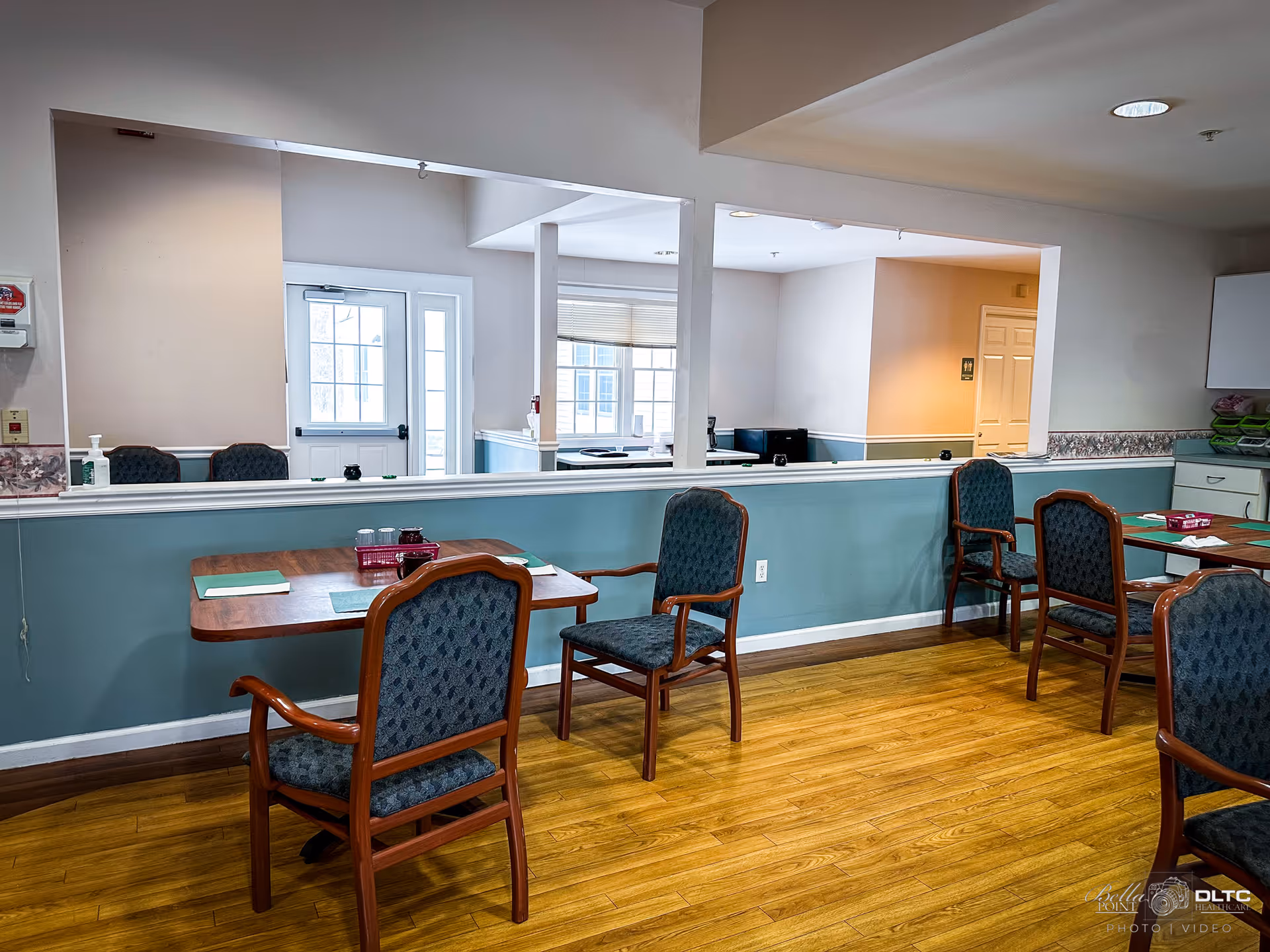 A dining area with wooden tables and upholstered chairs arranged on a wooden floor. The walls are painted light blue with a white trim, and there is a large open window-like cutout in the wall showing an adjacent room with windows and a door. The room is well-lit with natural light coming through the windows.