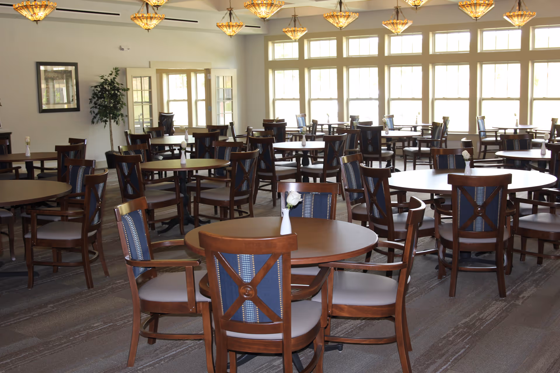 Bright dining room with multiple round wooden tables and chairs arranged throughout and large windows along the back wall.