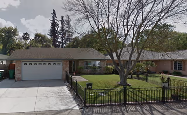 Single-story residential building with a two-car garage, a well-maintained front lawn enclosed by a black metal fence, and a large tree in the yard. The sky is partly cloudy and there are other houses visible on either side.