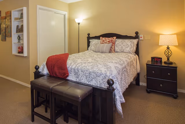 A warmly lit bedroom with a double bed dressed in patterned bedding, a bench at the foot, and a nightstand with a lamp and alarm clock.