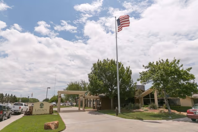 Front entrance of Legacy Rehabilitation And Living with a covered drive, a tall American flag, landscaping and a facility sign under a partly cloudy sky.