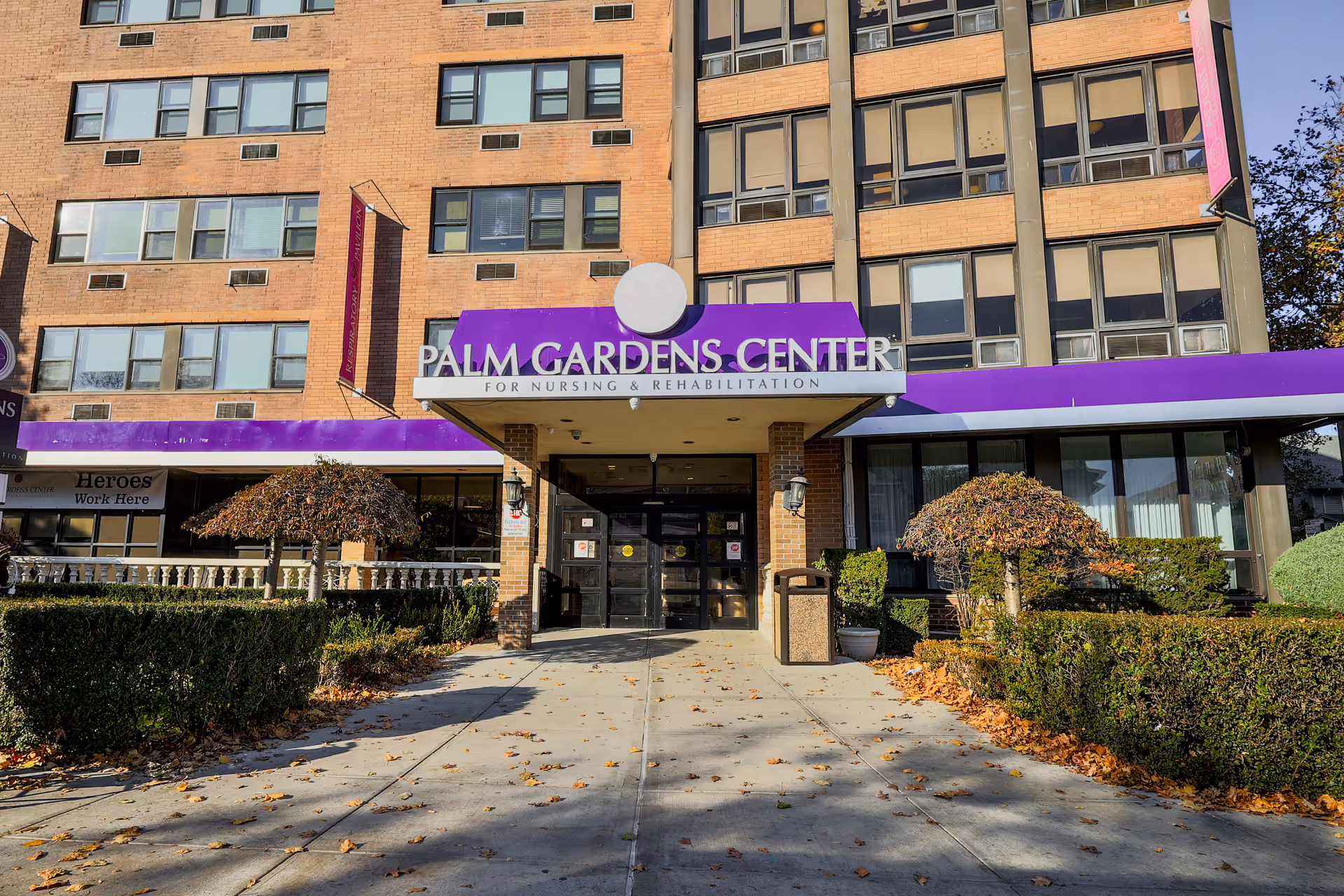 Front exterior view of Palm Gardens Center for Nursing & Rehabilitation, a multi-story brick building with a purple awning above the entrance. There are trimmed bushes and small trees along the walkway leading to the main doors.