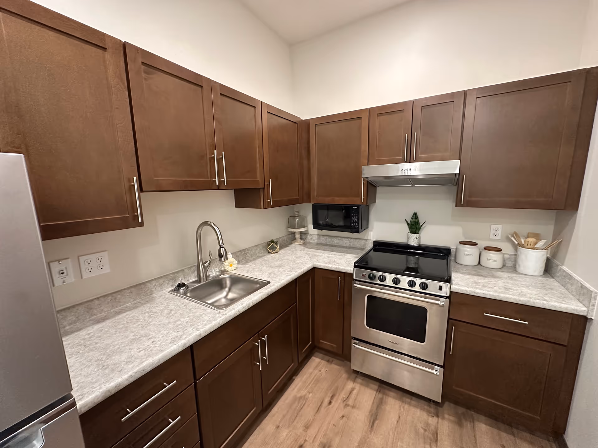 A modern kitchen with brown wooden cabinets, a stainless steel sink with a faucet, a stainless steel stove with an oven, a microwave mounted under the cabinet, and a refrigerator partially visible on the left. The countertops are light gray with a marble-like pattern, and there are small decorative items including a plant and kitchen canisters on the counter. The floor is wood with a light finish.