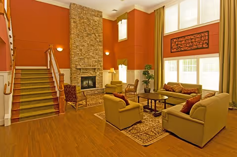 A spacious living room with high ceilings and large windows letting in natural light. The room features a stone fireplace in the center, surrounded by comfortable seating including sofas and armchairs arranged around a wooden coffee table on a patterned rug. There is a staircase with a green carpet runner on the left side, and the walls are painted a warm orange color with white trim. Green curtains frame the windows, and a decorative wall piece hangs above the windows.