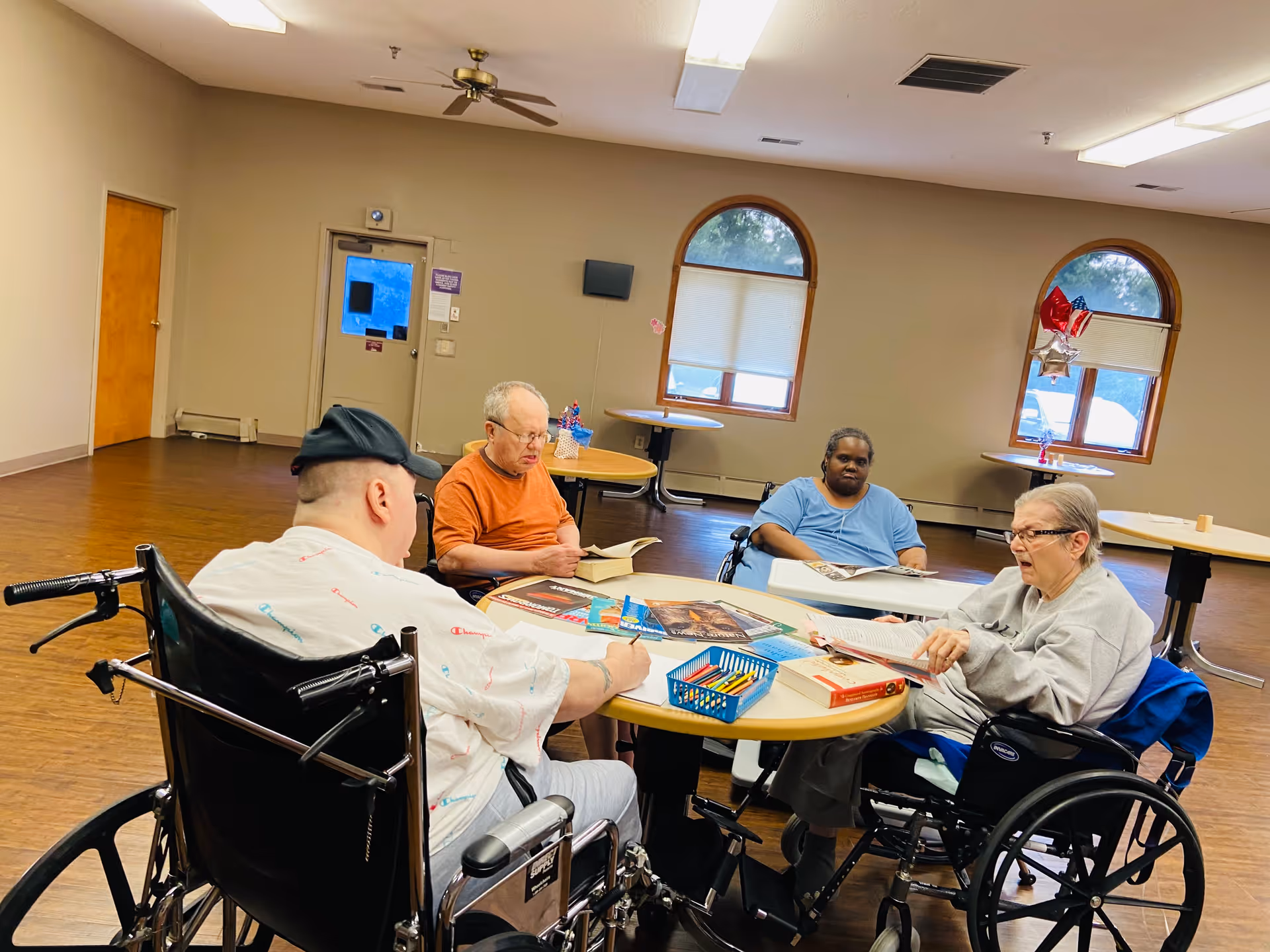Four elderly individuals, three in wheelchairs, sitting around a round table in a spacious room with wooden floors and beige walls. They are engaged in reading and writing activities with books, papers, and colored pencils on the table. The room has arched windows with blinds and some balloons tied to one window.