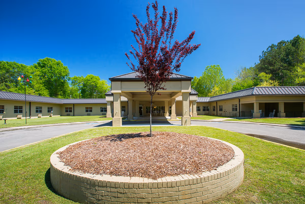 Front exterior view of Moundville Health and Rehabilitation, LLC building with a circular brick planter containing a small tree in the foreground, surrounded by a driveway and green lawn under a clear blue sky.