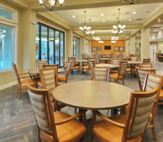A spacious dining room with multiple round and rectangular wooden tables surrounded by cushioned chairs. Large windows line one side of the room, allowing natural light to fill the space. The ceiling features multiple chandeliers, and the walls are decorated with framed pictures and a mounted television.
