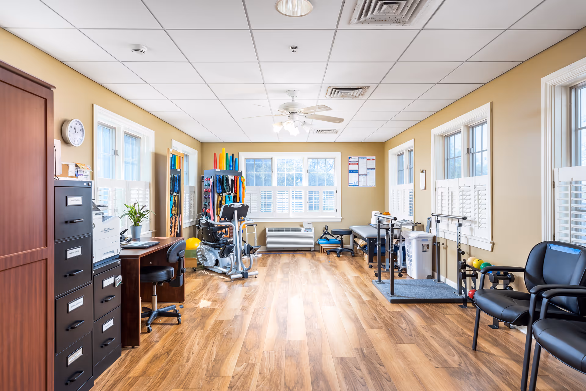 A bright and spacious therapy or rehabilitation room with wood flooring, beige walls, and multiple windows with white shutters. The room contains exercise equipment, therapy tools, a desk with a chair, filing cabinets, and several black chairs along the right wall. There is a ceiling fan with lights and informational posters on the walls.