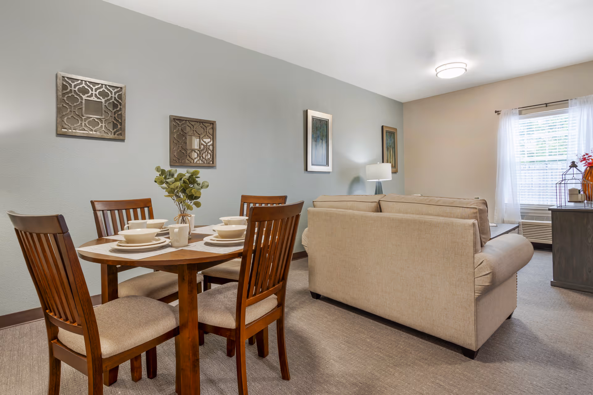 Bright furnished living and dining area with a wooden dining table set and a beige sofa in a senior living apartment.
